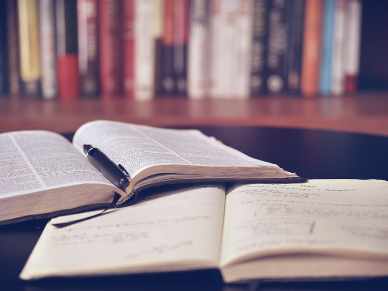 An open book and notebook on a table in front of a bookshelf