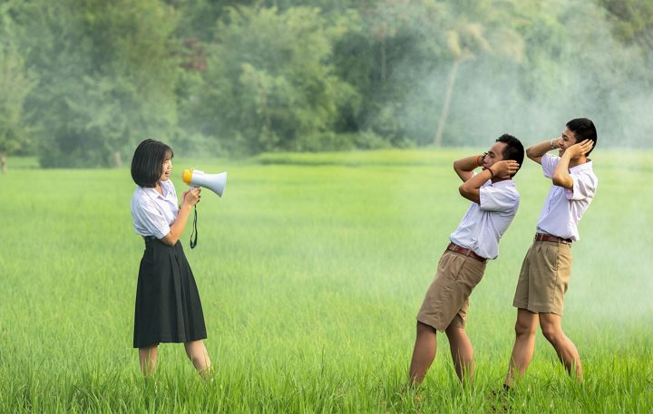 A student speaking through a megaphone in a meadow while two others cover their ears
